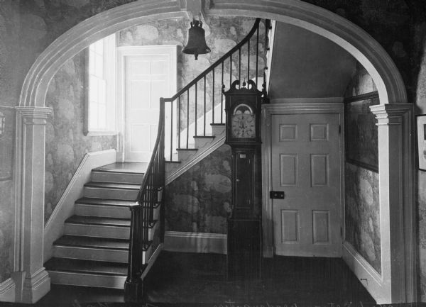 Interior view of the Morris-Jumel Mansion which served, at different times, as headquarters to both forces during the Revolutionary war.  This view of the stairway features an arched entryway with a bell hanging from the top, and a grandfather clock.