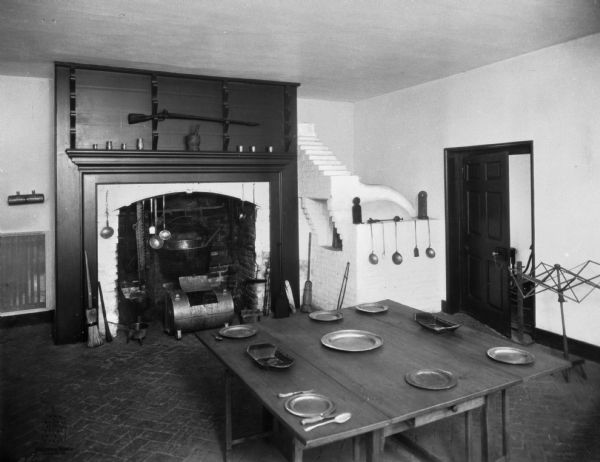 Interior of farmhouse kitchen featuring a fireplace for cooking, an adjacent brick oven, and a table in the center of the room set with metal dishes. Cooking implements are hanging above the fireplace, and a rifle is mounted above the mantle.