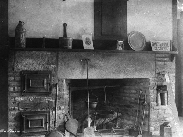 Cooking fireplace at the birthplace of Daniel Webster, a house that was built in 1780 and restored in 1913. There are oven doors built into the left side of the fireplace, hanging pots in the fireplace, and tools and cooking related items. A sign on the mantel reads: "Do Not Handle The Souvenirs."