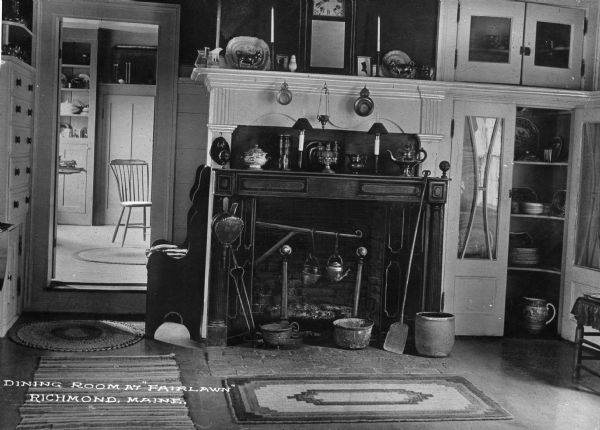 The Fairlawn home's colonial dining room. Features area rugs, a fireplace with hanging kettles, fireplace tools, displayed plates and figurines and a built in cabinet for dishes. Caption reads: "Dining Room at 'Fairlawn' Richmond, Maine."