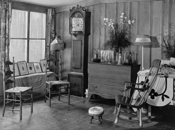 View of a parlor in the Three Villages Tea House. The room features a sitting area, a grandfather clock, lamps, a flower arrangement and displayed vases and a row of framed crests.