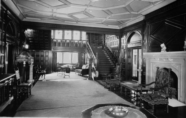 Interior view of the reception hall at Ladywood School. Features a fireplace in the foreground, a sitting area near the window in the back, extensive wood paneling throughout the room, carved wooden furnishings and a grand staircase leading to the second floor.