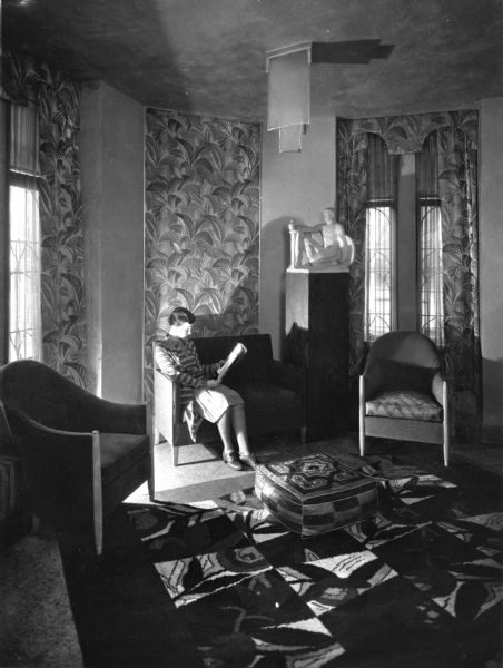 Interior view of sitting room with a tall ceiling. The room is decorated in art deco style with a light fixture, upholstered chairs, patterned carpet, patterned curtains, an embroidered ottoman and a sculpture, "Oasis - The Panhellenic." A woman, lit by the sun, sits on a sofa reading.