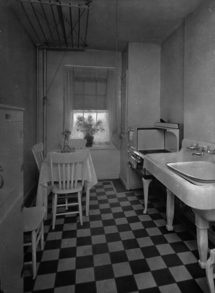 View of the kitchen at Amalgamated Clothing Workers Cooperative apartments. The narrow room features a checker-tiled floor, a small table and chairs, a flower arrangement, potted plant on the window sill, gas stove, ice box, large sink and retracted drying rack at the ceiling.