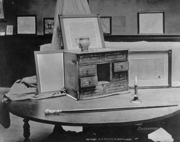 Chest brought to America on the Mayflower. Features a ceramic cup atop it while a walking cane and candle holder sit on the table next to the chest.