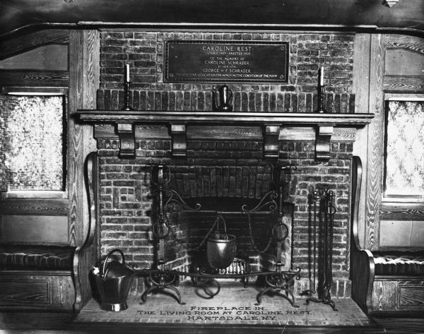 View of fireplace with tools, decorative stands, an ash can and flanking benches below windows. Above the fireplace an engraved dedication reads: "Caroline Rest: Founded 1907 - Erected 1908. To the memory of Caroline Schrader by her son George H F Schrader." Caption reads: "Fireplace in the Living Room at Croline Rest, Hartsdale, N.Y."