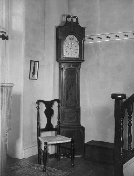 View of a grandfather clock at the foot of a set of stairs, featuring the end railing of the staircase, a chair, and a framed silhouette of a man hanging on the wall.