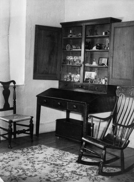View of combination desk and cabinet in a corner with two chairs on either side. The cabinet doors are open showing glassware and ceramics inside.