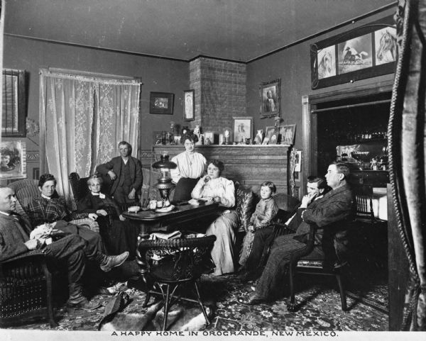 View of a family relaxing in the living room of their home. Features men, women, and children sitting around a table. Caption reads: "A Happy Home in Orogrande, New Mexico."