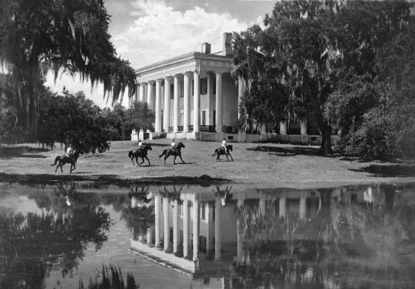 View of four horses and riders galloping across the front lawn of a southern mansion, next to a body of water that reflects the scene. A large, white mansion with pillars is centered at the back of the photograph and large trees trailing Spanish moss flank the building.
