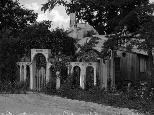 Exterior view of a small building isolated in the woods. In the foreground is a gateway of arches to a patio and oratory overgrown with ivy.
