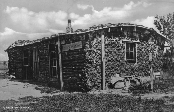 View of sod house in short grass country. Features rectangular building with a curved roof and windows.
