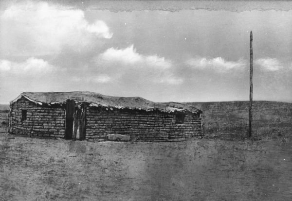 View of first house built in Dodge City. The sod building has a roof, window openings and a door. The surrounding landscape is barren.