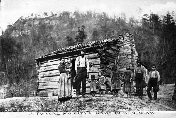 Group portrait of a man, a woman and seven children standing in front of a log cabin. The caption reads: "A Typical Mountain Home in Kentucky."