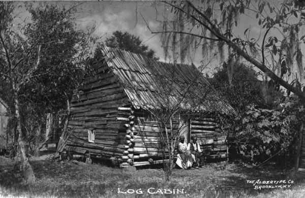View of a dilapidated log cabin with among trees. A man and woman are sitting on the front step. Caption reads: "Log Cabin."