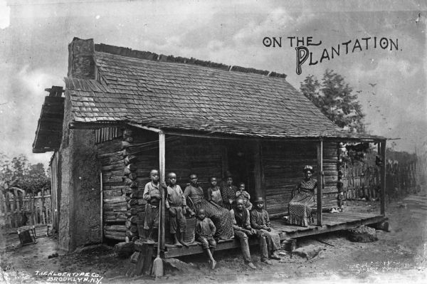 View of a group of three women and seven children sitting on the porch of a log cabin. The caption at top right corner reads: "On The Plantation."