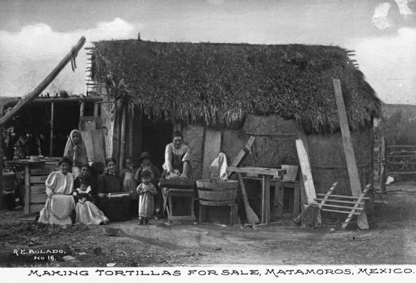 View of four women and five children posing outside of a thatch-roofed home. Work tables and a barrel tub are gathered in front of the building and one woman is standing while grinding masa for corn tortillas with a mano and a metate. Caption at bottom reads: "Making Tortillas For Sale, Matamoros, Mexico."