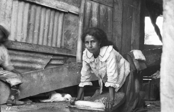 View of a woman kneeling and grinding corn by hand with the traditional mortar and pestle, the mano and metate. On the left a child is partially visible siting on a low beam, with a dog sitting underneath it.