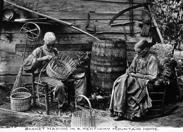View of old man making baskets and an old woman sewing outside of a log cabin. Other handmade wooden items appear throughout the image and tools hang on the cabin wall. Caption at bottom reads: "Basket Making in a Kentucky Mountain Home."