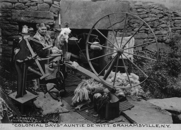 View of woman sitting with spinning wheel, bulk wool and a yarn winder by a stone wall with a cooking fireplace in it. Plants grow on the ground nearby, the setting may be a historical reenactment in the ruin of a colonial house. The caption reads: "'Colonial Days' Auntie Dewitt, Grahamsville, N.Y."