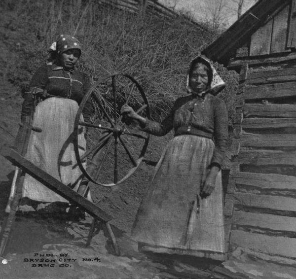 View of two women with a spinning wheel standing in front of a small wooden building.