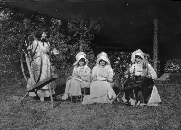 View of four women on a lawn wearing dresses and bonnets, posing with wool and flax spinning wheels.