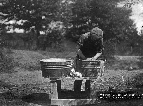 View of a person doing laundry in metal tubs on a bench outside.  The caption reads: "'The Hand Laundry' Lake Huntington, N.Y."