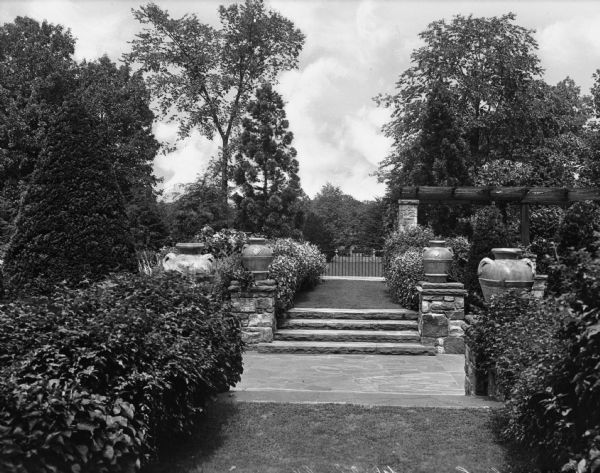 View of pathway through an elaborate garden on the Walhall Estate. Features large ceramic vases, a trellis and a metal fence as well as a stone pathway and staircase in the center of the garden.
