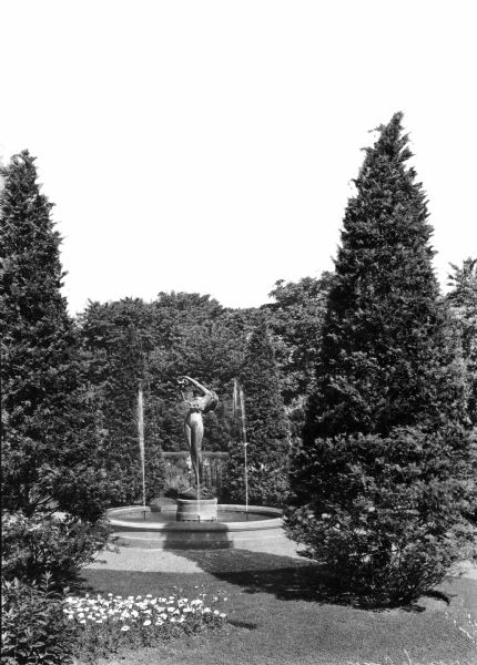 View of fountain in a garden of the Walhall Estate. The fountain has a sculpture of a flower-adorned woman, and the garden features evergreen shrubs along with a bed of daisies.