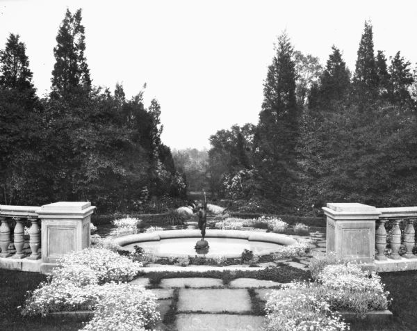 View of Wavery Garden, featuring a fountain with a sculpture of a boy raising a cup to the sky, carved stone balustrades, tufts of flowers and a background of trees.