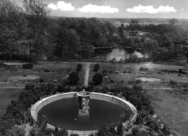 Elevated view of a formal garden, a fountain and a pond bordered by woods.