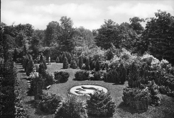 Raised view of a garden at Ladywood School, including a statue, a flower-ringed sunken fountain, a stone bench and trees and bushes.