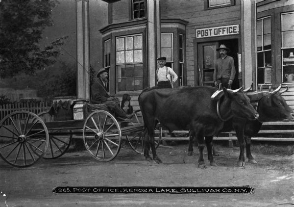View of two large oxen pulling a wagon stopped in front of a post office.  On the steps in front of the building two men stand looking towards the street.  Farther along the steps sits a dog. Caption at bottom reads: "965. Post Ofice, Kenoza Lake, Sullivan Co. NY"