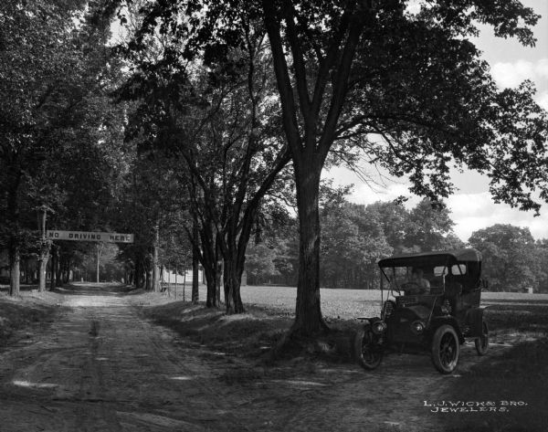 A dirt road through the woods with a man in the driver's seat of a car in the right foreground. A sign is hanging above the road that reads: "No Driving Here."
