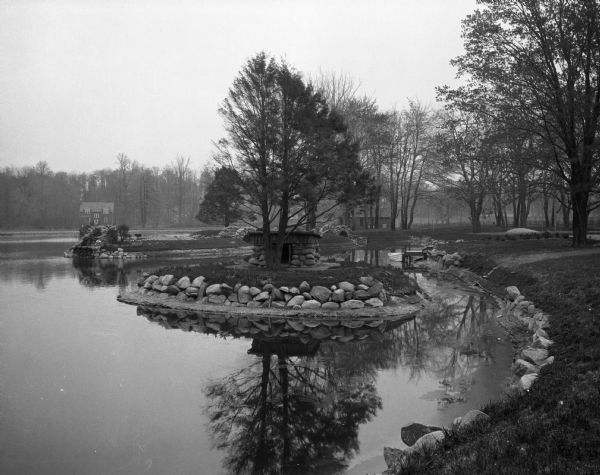 Park with stone-lined waterways, surrounded by woods.  In the middle of the image is a small island with a rustic stone hut and in the background are stone bridges and steps to the water.