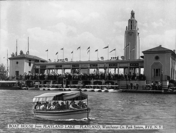 View across water toward s large boathouse with a crowd of people in front, and an excursion boat in the foreground on Playland lake. Caption reads: "Roat House from Playland Lake — Playland, Westchester Co. Park System, Rye, N.Y."