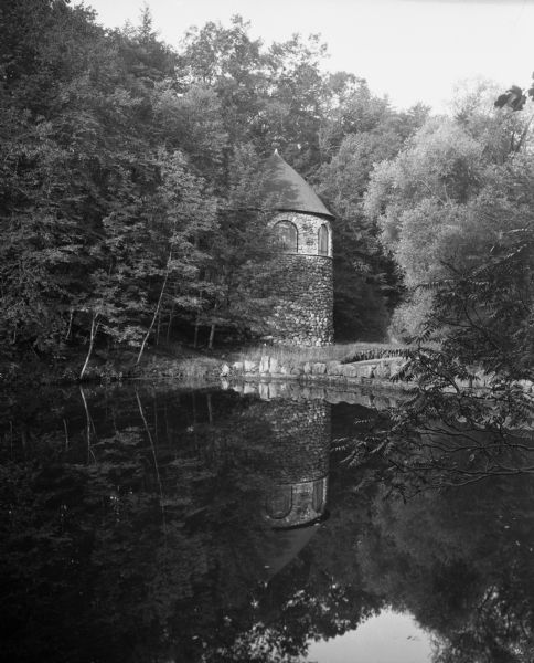 Small round stone building in the woods reflected in a pond or lake.