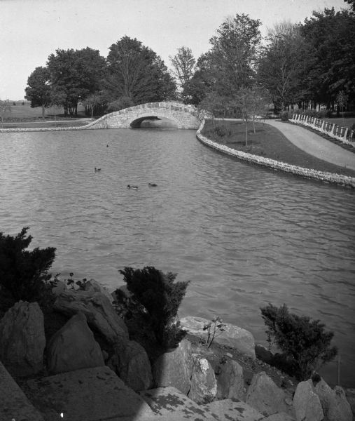 View of a stone lined lake in a park with trees, a paved path, a stone bridge and ducks.