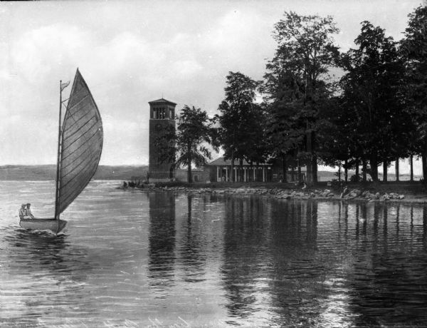 Two sailors in a sailboat on Lake Chautauqna with the Lewis Miller Memorial bell tower on the point in the background.
