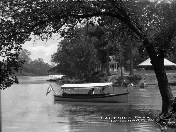 Two men in a motorized boat with an awning cruise past a park with a tent and a gazebo. Caption reads: "Lakeside Park, Carthage, MO."