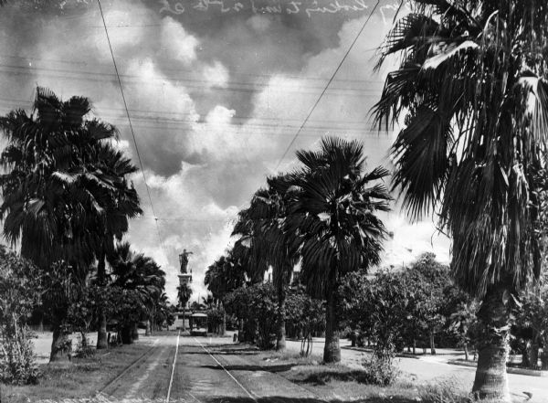View looking down Broadway, flanked by palm trees, at the Rosenberg Monument. Street car tracks lead to the monument and a trolley is visible in the distance.