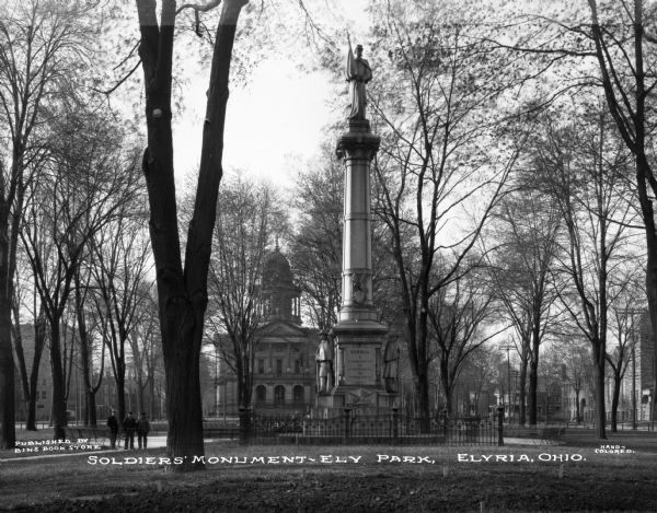 Tall cylindrical Civil War memorial with multiple sculptures in Ely Square in downtown. A wrought iron fence surrounds the monument, trees dot the square and the Lorain County courthouse with its original dome is in the background. Caption reads: "Soldiers' Monuments — Ely Park, Elyria, Ohio."