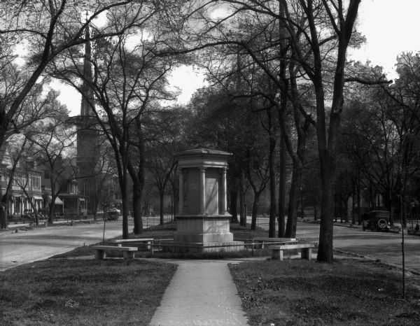 Small monument to southern poets surrounded by benches in a median park on Greene Street. Trees line the street and large stone buildings are in the background.