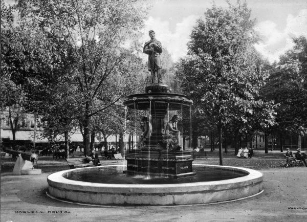 Civil War memorial statue and fountain in a town park with a cannon in the background. Parkgoers relax on the surrounding benches in the shade of trees.