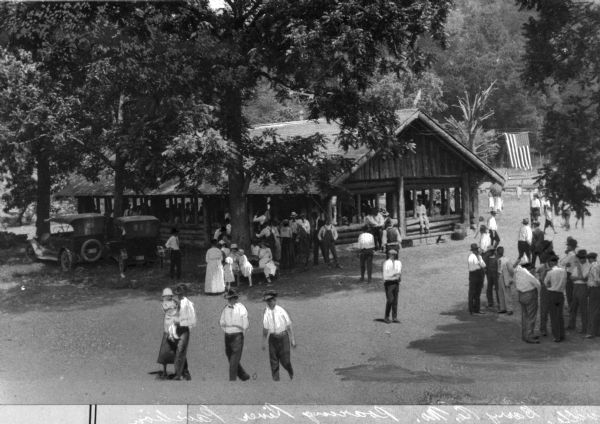 Elevated view of children and adults relaxing and socializing at a camp in the woods. The camp has a large, open, log building, and a flag is hanging in the background. Two cars are parked nearby.