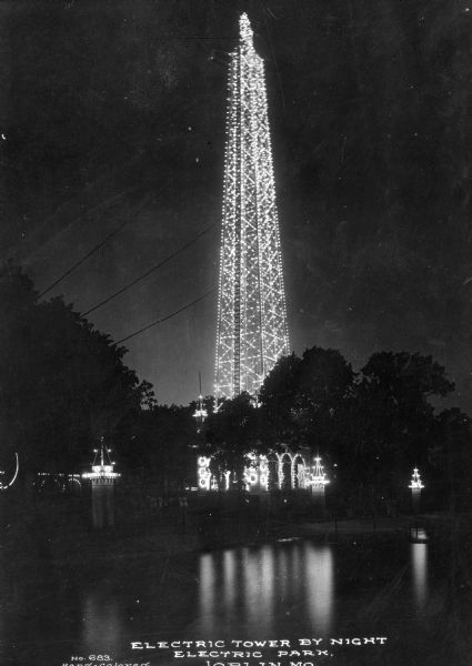 View across water toward a tall, lighted, electric tower at night with trees and lighted posts surrounding it among trees. Caption reads, "Electric Tower by Night, Electric Park, Joplin, MO."