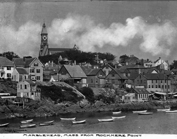 Elevated view of village homes on the shores of the bay with docked fishing boats in the foreground. Caption reads, "Marblehead, Mass from Rockmere Point."
