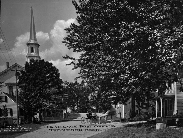 View down dirt road toward a small horse-drawn carriage and surrounding town buildings, including the post office and a church with a steeple. Caption reads, "The Village Post Office, Thompson, Conn."