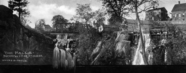 View toward two waterfalls flowing over rocks into a ravine. One waterfall is held back by a dam and the other is traversed by a railroad bridge. Trees and houses line the edges of the ravine.  Caption reads, "The Falls - Norwich, Conn."