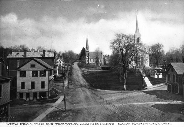 Elevated view of town's central dirt road from the railroad trestle looking north toward large wooden buildings and two churches. Caption reads, "View from the R.R. Trestle, looking north. East Hampton, Conn."
